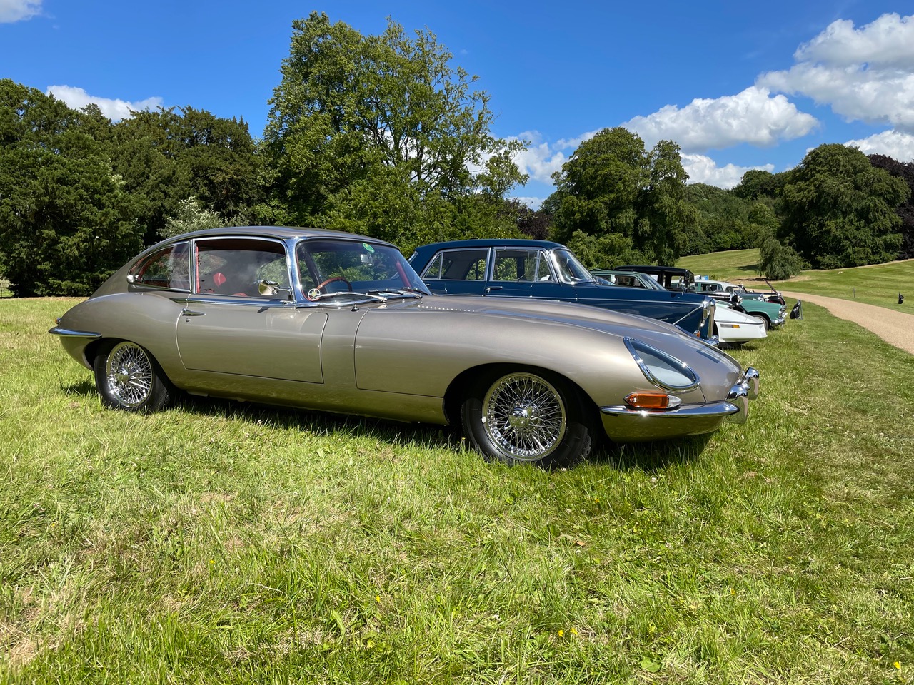 Front three-quarter view of a Golden Sand 1967 Jaguar E-Type Series 1 4.2 2+2 registered TUK 129F on grass at a classic car show, its glass headlight covers and chrome wire wheels visible