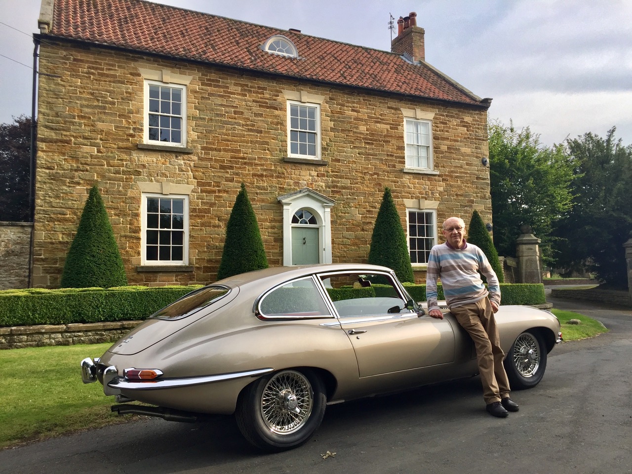 Stephen Ruddick standing beside his Golden Sand Jaguar E-Type Series 1 4.2 2+2, seen from the rear three-quarter in front of a stone house