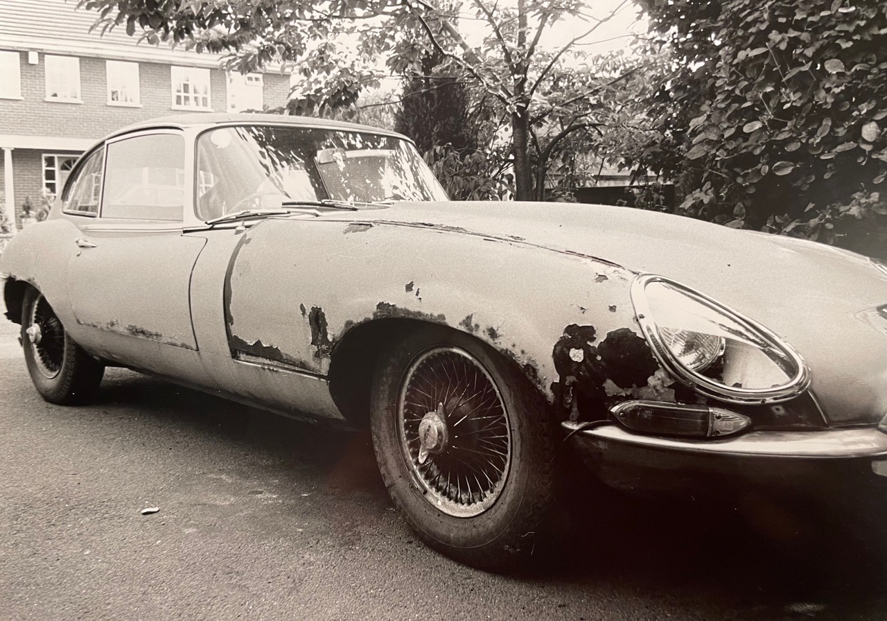 A Jaguar E-Type before restoration, showing heavy corrosion through the front wing, sill and headlight surround, and paint lifting along the body