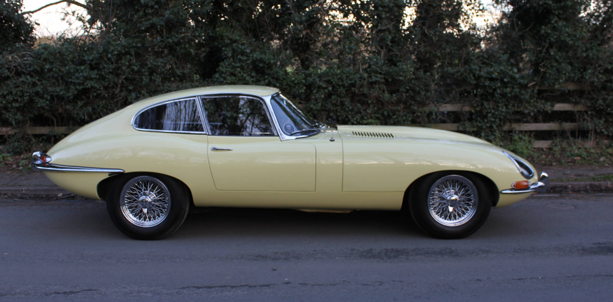 Side profile of a Primrose Series 1 E-Type fixed-head coupe on a road in front of a hedge